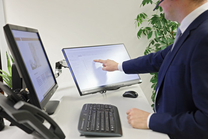 Aufgaben des Ministeriums Eine männliche Person stehend vor einem Schreibtisch mit zwei Monitoren, einem Telefon und einer Tastatur. Männliche Person bedient einen Touch-Bildschirm